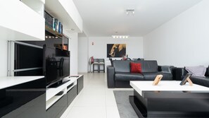 Een moderne woonkamer met een flatscreen televisie, een zwarte leren bank, een salontafel, en een eethoek met stoelen en een kleine tafel.