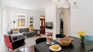 Een moderne woonkamer met een grijze bank, zwarte fauteuils, een houten eettafel met een schaal fruit, en een tv die aan de muur is bevestigd.