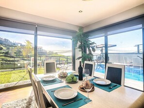 A dining area with a table set for two, large windows offering a view of a pool and greenery, and a ceiling light fixture.