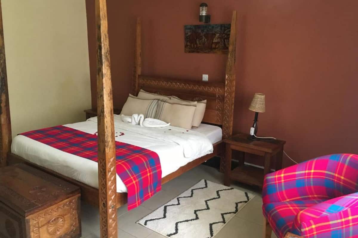 A four-poster bed with a plaid bedspread, a nightstand with a lamp, a chair with a plaid cushion, and a painting on the wall.