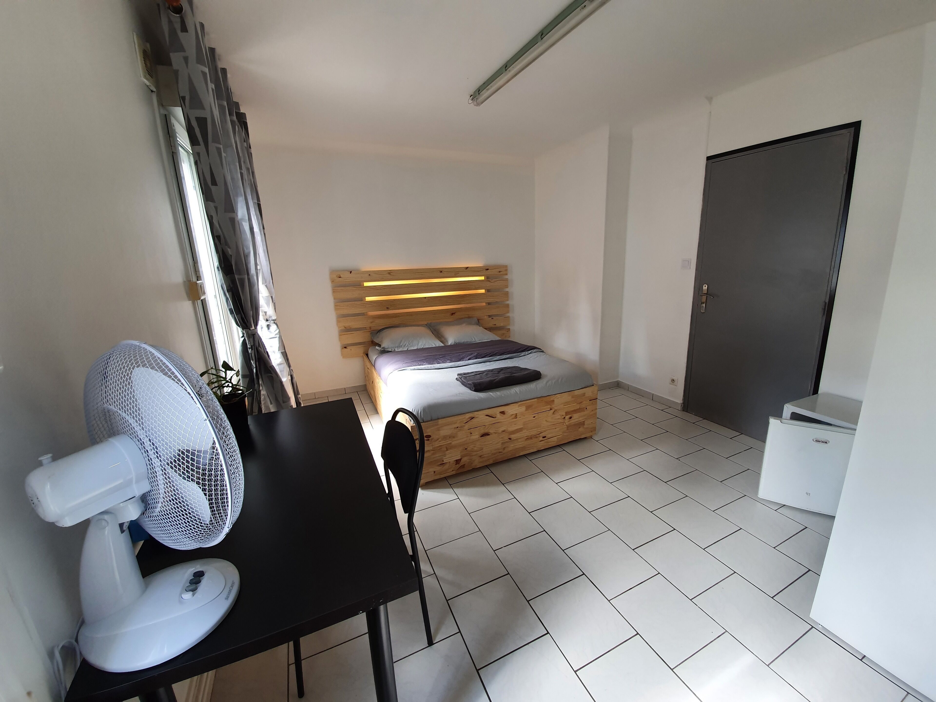 A bedroom with a bed, a desk with a fan, a door, and a small white cabinet.