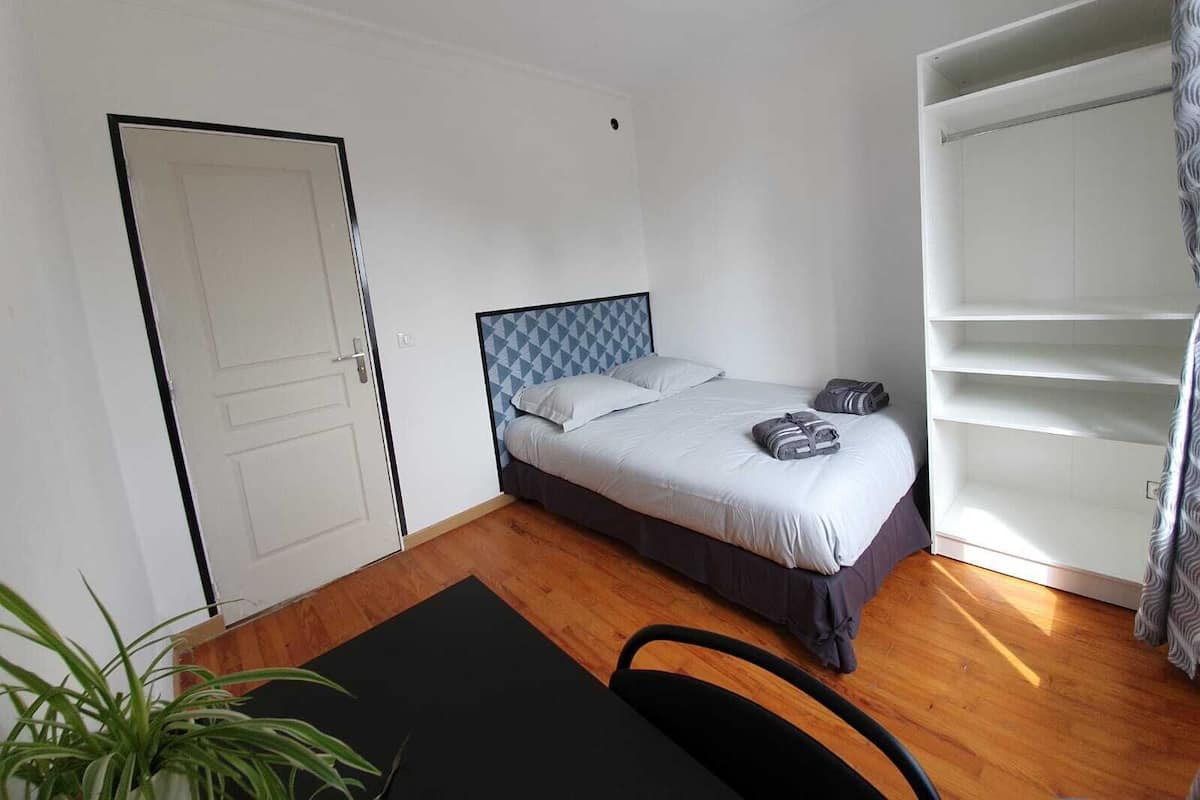 A small, clean room with a bed, a desk, and a chair. There is a door and a wardrobe with shelves.