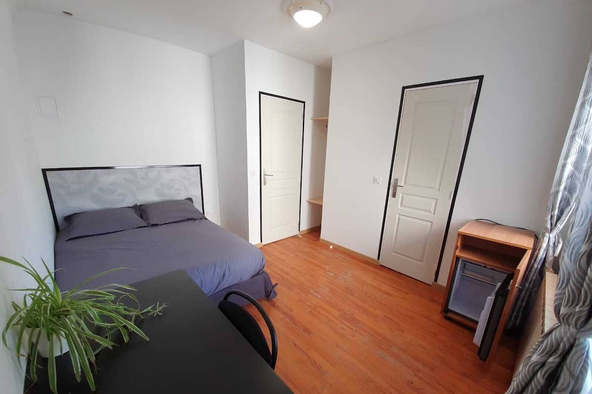 A bedroom with a bed, a desk, a chair, a potted plant, and two doors.