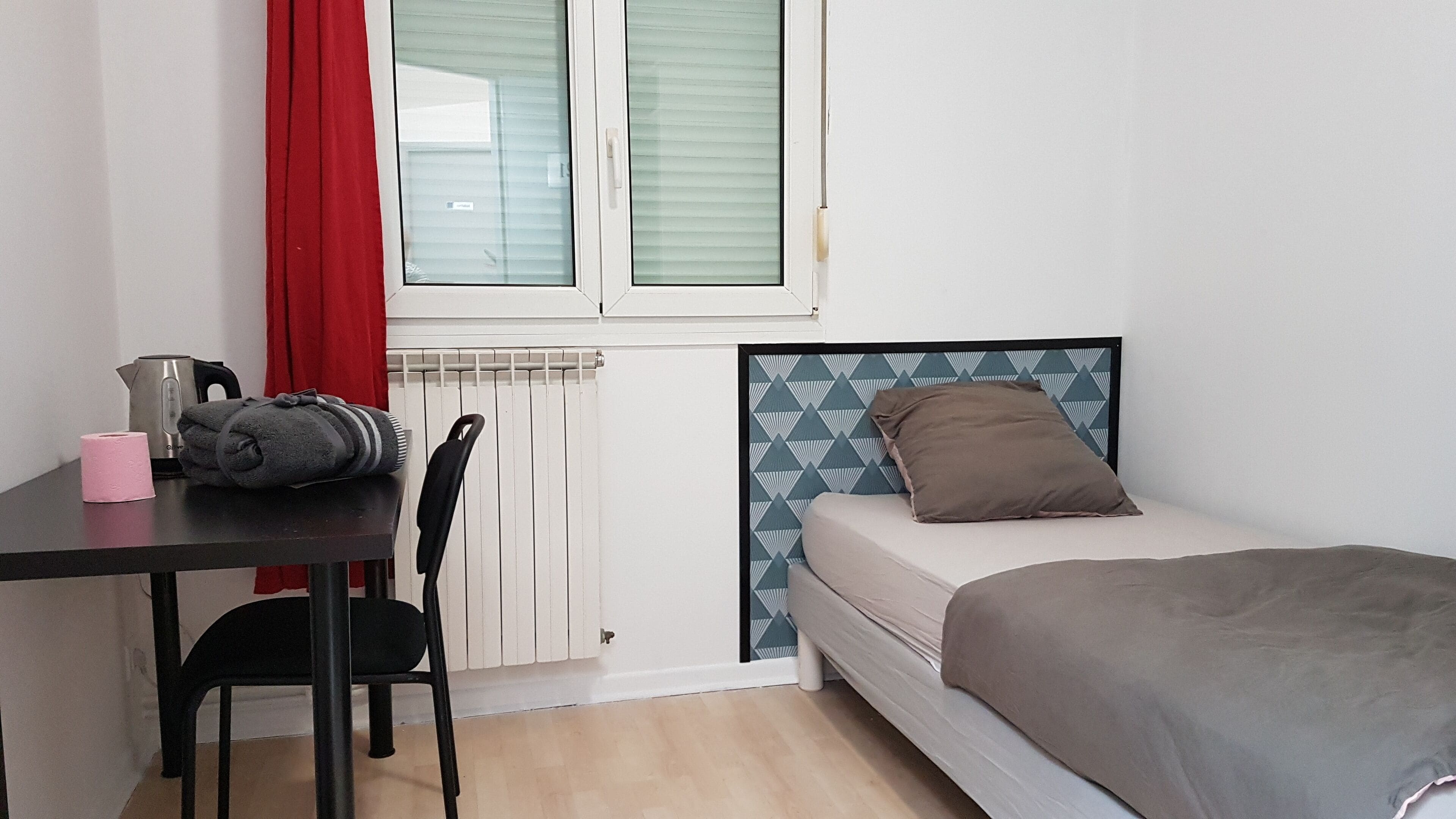 Une petite pièce propre et bien rangée, avec un lit, une chaise, une table sur laquelle se trouvent une bouilloire et un rouleau de papier toilette, et une fenêtre avec des stores.