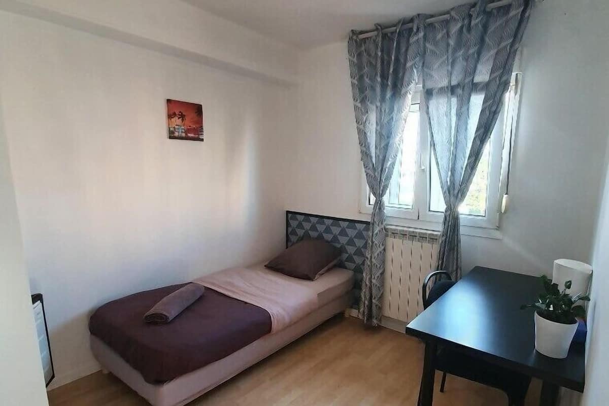 A small, single-bed room with a wooden floor, a small table, and a window with curtains.