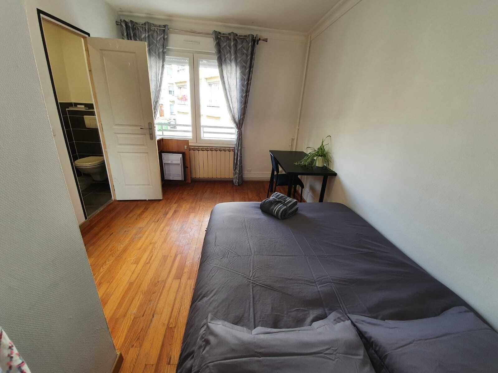 Une petite pièce propre et bien rangée, avec un lit, un bureau et une fenêtre avec des rideaux.