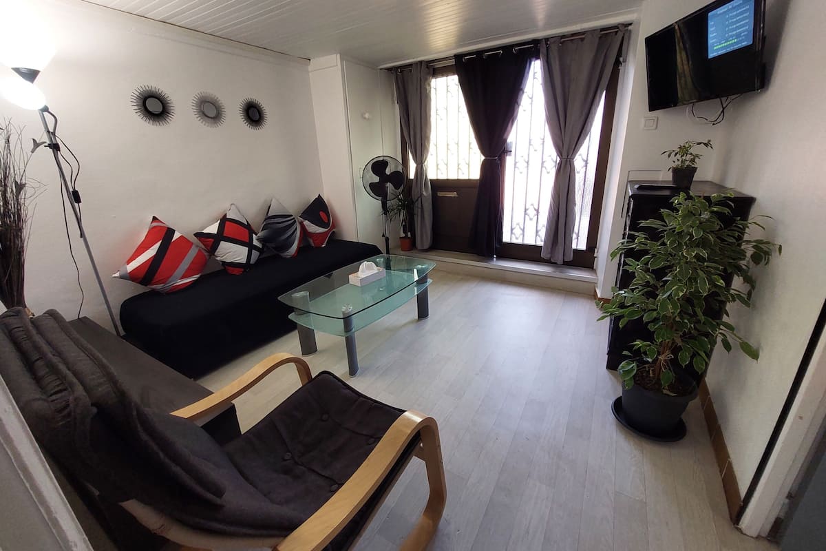 A living room with a black sofa, a glass coffee table, a wooden chair, a TV mounted on the wall, and a potted plant.