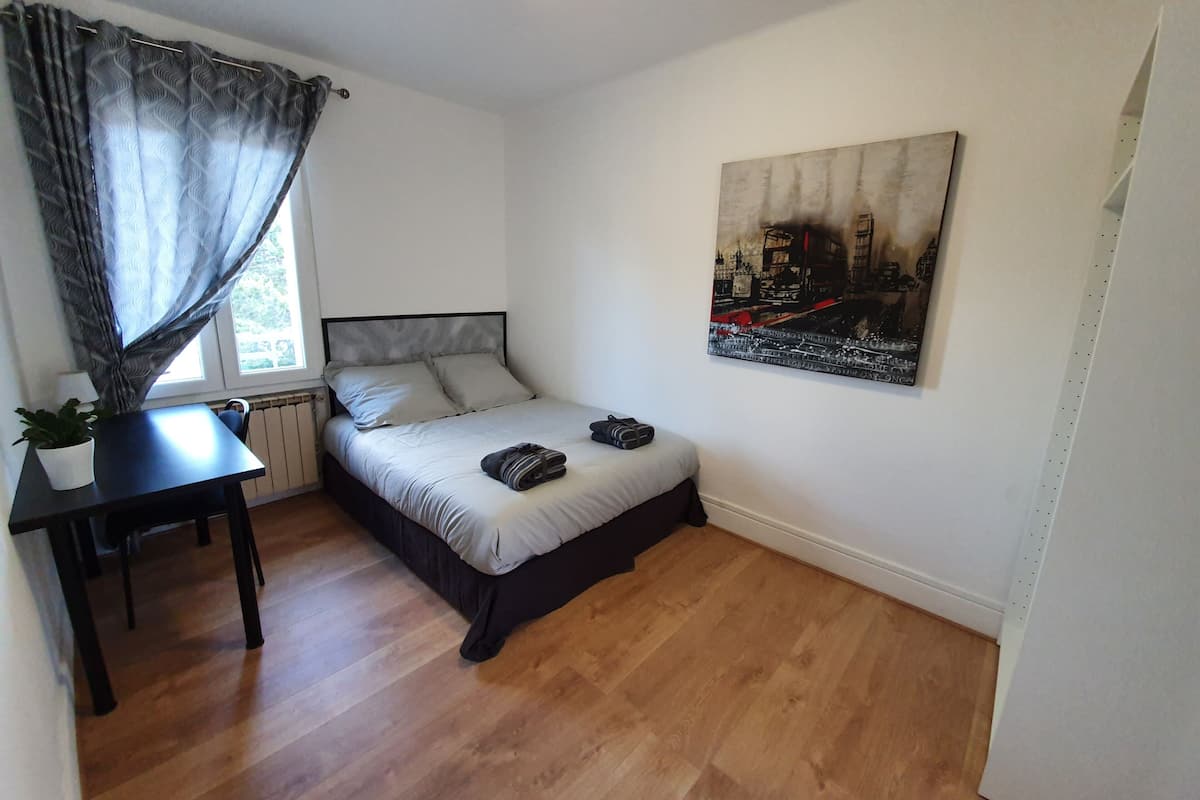 A bedroom with a bed, a desk, a chair, a potted plant, and a painting on the wall.