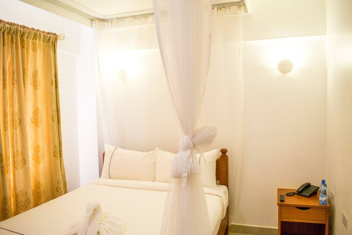 A hotel room with a bed, a nightstand with a phone and a bottle, and a curtain.