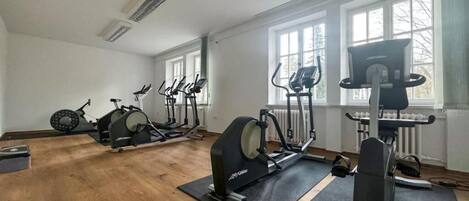 Un gimnasio con diversas máquinas de ejercicio, amplios ventanales y suelo de madera.