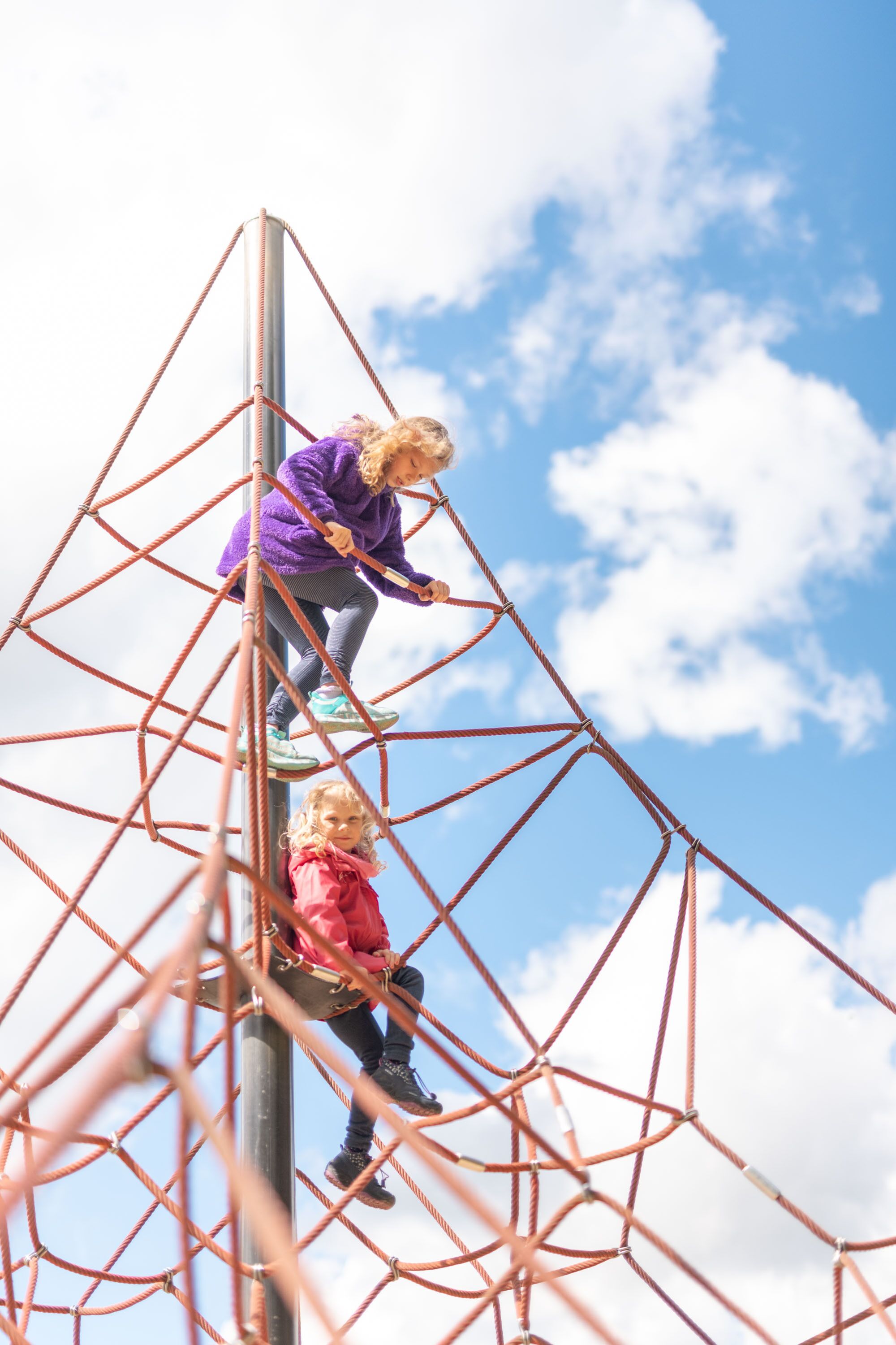 Two children climbing a rope ladder structure against a clear sky.