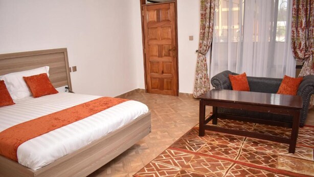 Une chambre à coucher avec un lit, une tête de lit en bois, un tapis à motifs, un canapé avec des coussins orange et une porte en bois.