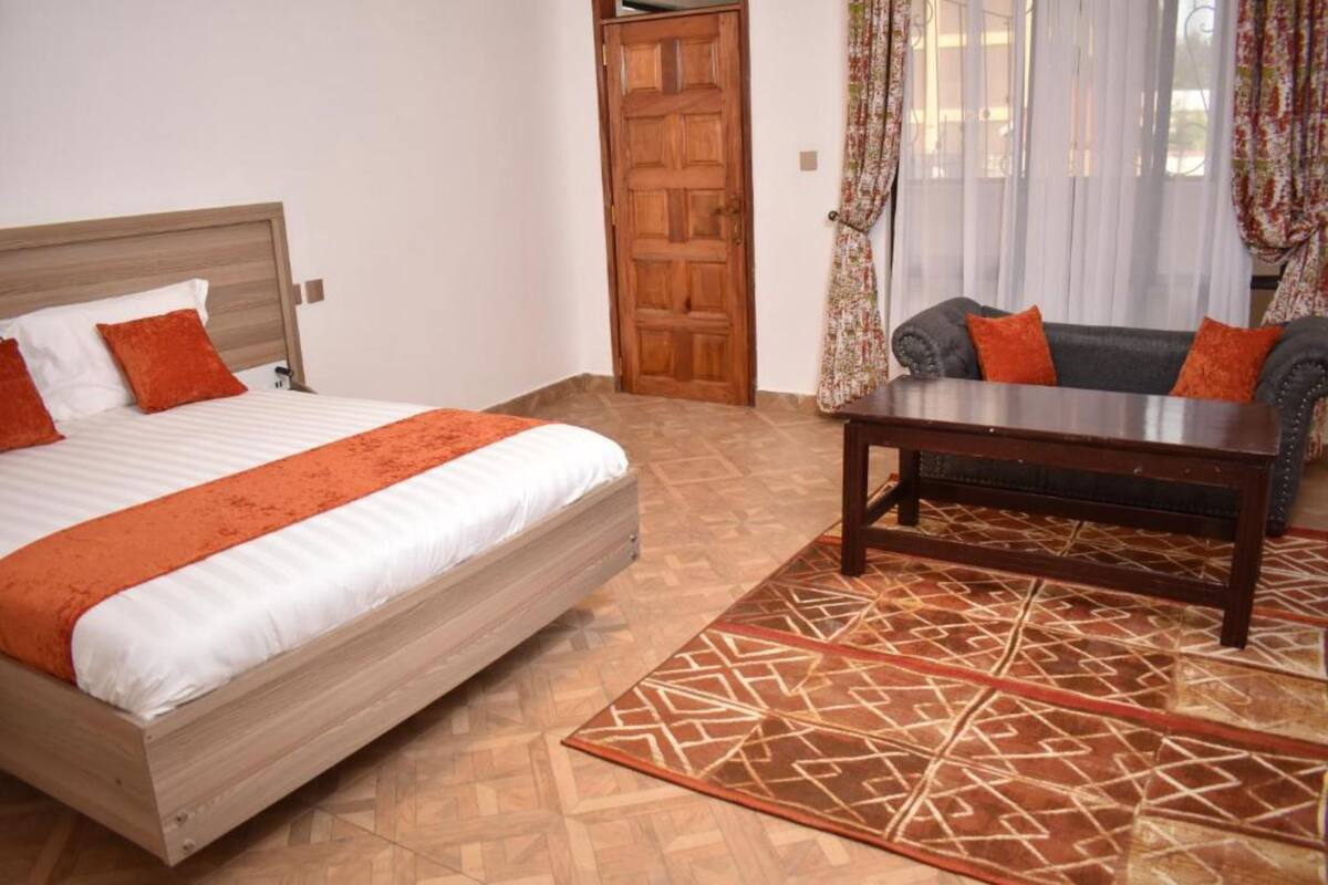 Une chambre à coucher avec un lit, une tête de lit en bois, un tapis à motifs, un canapé avec des coussins orange et une porte en bois.
