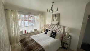 Un dormitorio con cama, candelabro, ventana con cortinas, mesita de noche y una obra de arte en la pared.