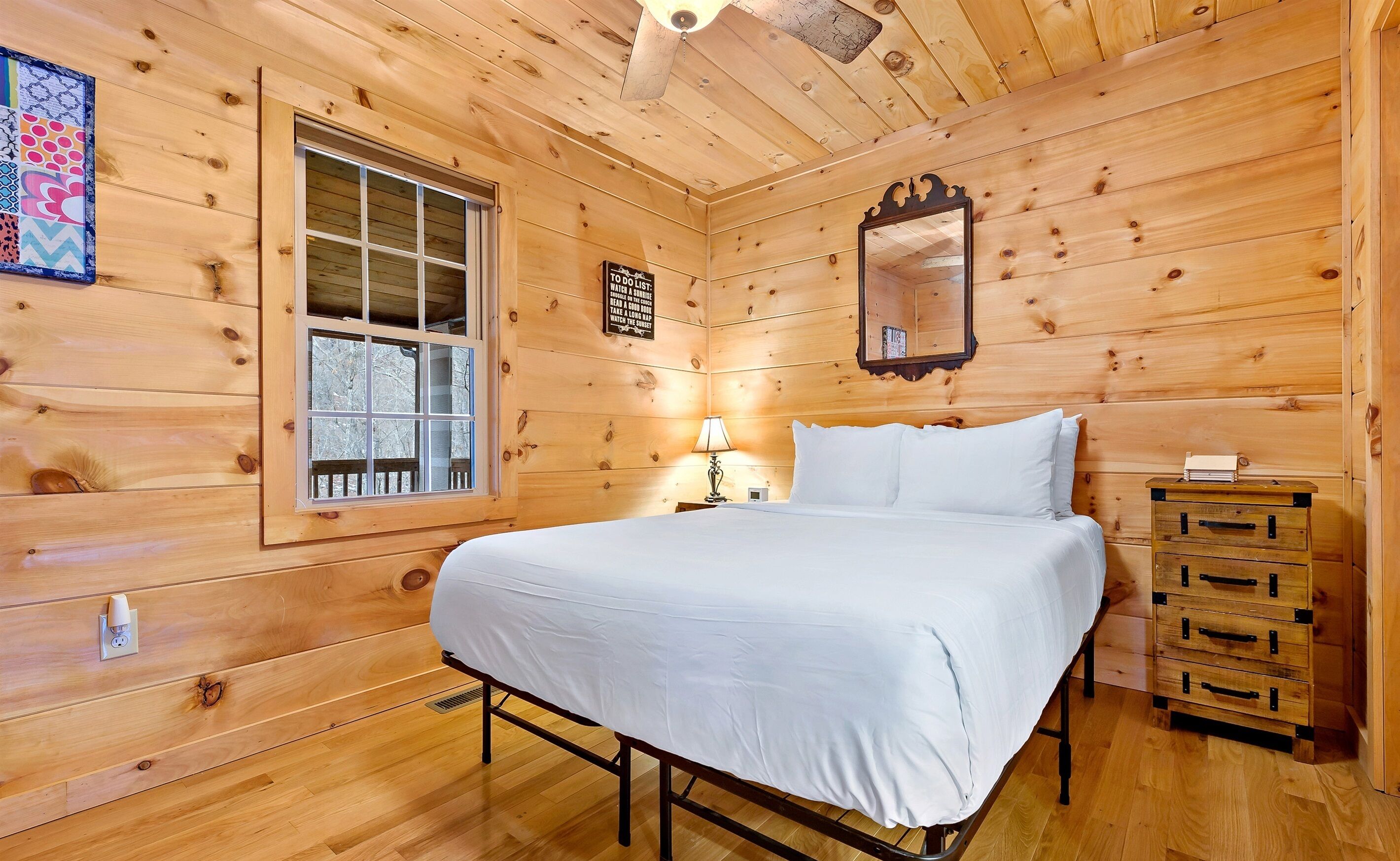 Una habitación de cabaña de madera con una cama, un ventanal, un espejo, una lámpara y un aparador.