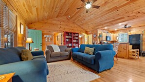 A cozy cabin interior with a wooden ceiling, a living room with a blue sofa and armchairs, a bookshelf, and a kitchenette. - Hot Tub, Mirror Lake Access, Log Cabin - Hawks Terrace by Carolina Properties (Lake Lure)