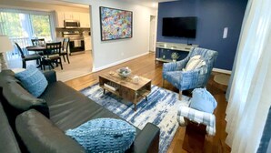 A modern living room with a dark sofa, a blue armchair, and a coffee table. There is a kitchen in the background with a dining area.