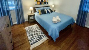 A bedroom with a bed, a ceiling fan, a nightstand with a lamp, a rug, and a painting on the wall.