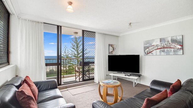 Ein modernes Wohnzimmer mit großem Fernseher, Ledersofas, einem kleinen runden Tisch und einem Balkon mit Meerblick.