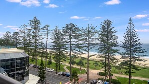 Blick auf einen Strand mit hohen Bäumen, einem Parkplatz und einem Gebäude mit Glasfassade.