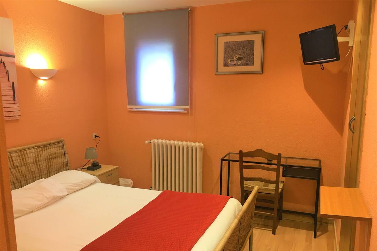 Une chambre d’hôtel équipée d’un lit, d’un bureau, d’une chaise, d’un radiateur, d’un téléviseur fixé au mur et d’une fenêtre avec des stores.