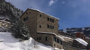 A stone building with multiple windows and a sloped roof, surrounded by snow-covered terrain and trees. - Hotel Roc de St Miquel (Soldeu)
