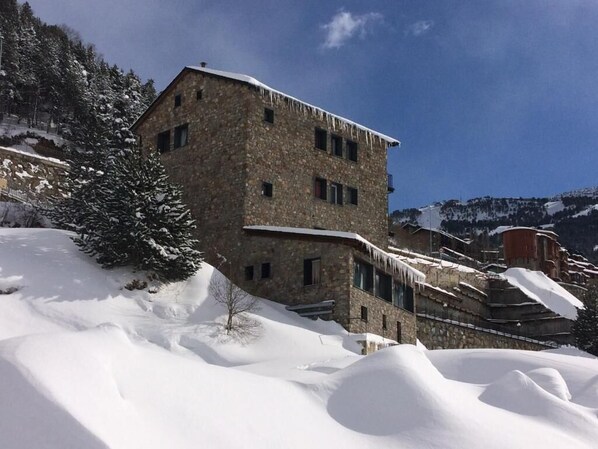 A stone building with multiple windows and a sloped roof, surrounded by snow-covered terrain and trees. - Hotel Roc de St Miquel (Soldeu)
