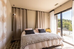 Quarto com cama grande, cortinas, abajur e vista para o verde lá fora.