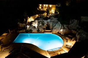 A swimming pool with a curved edge, surrounded by a rocky landscape and a building with multiple windows and lights. - Gîte Laumaga I 4 pers. I Spa privé I Domaine le C4 (Casteljau)