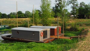 Zwei schwimmende Strukturen mit Deck und Rettungsboje, verbunden durch einen Holzsteg, inmitten einer natürlichen Landschaft mit Bäumen und einem Gebäude in der Ferne.