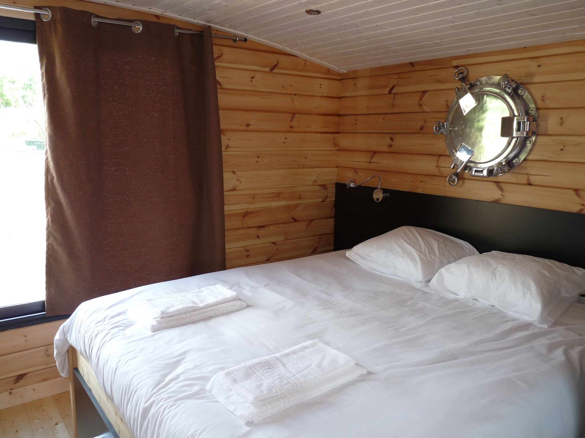 Ein Zimmer in einer Holzhütte mit einem Bett, weißen Bettwäsche, einem Fenster mit braunen Vorhängen und einem runden Spiegel an der Wand.