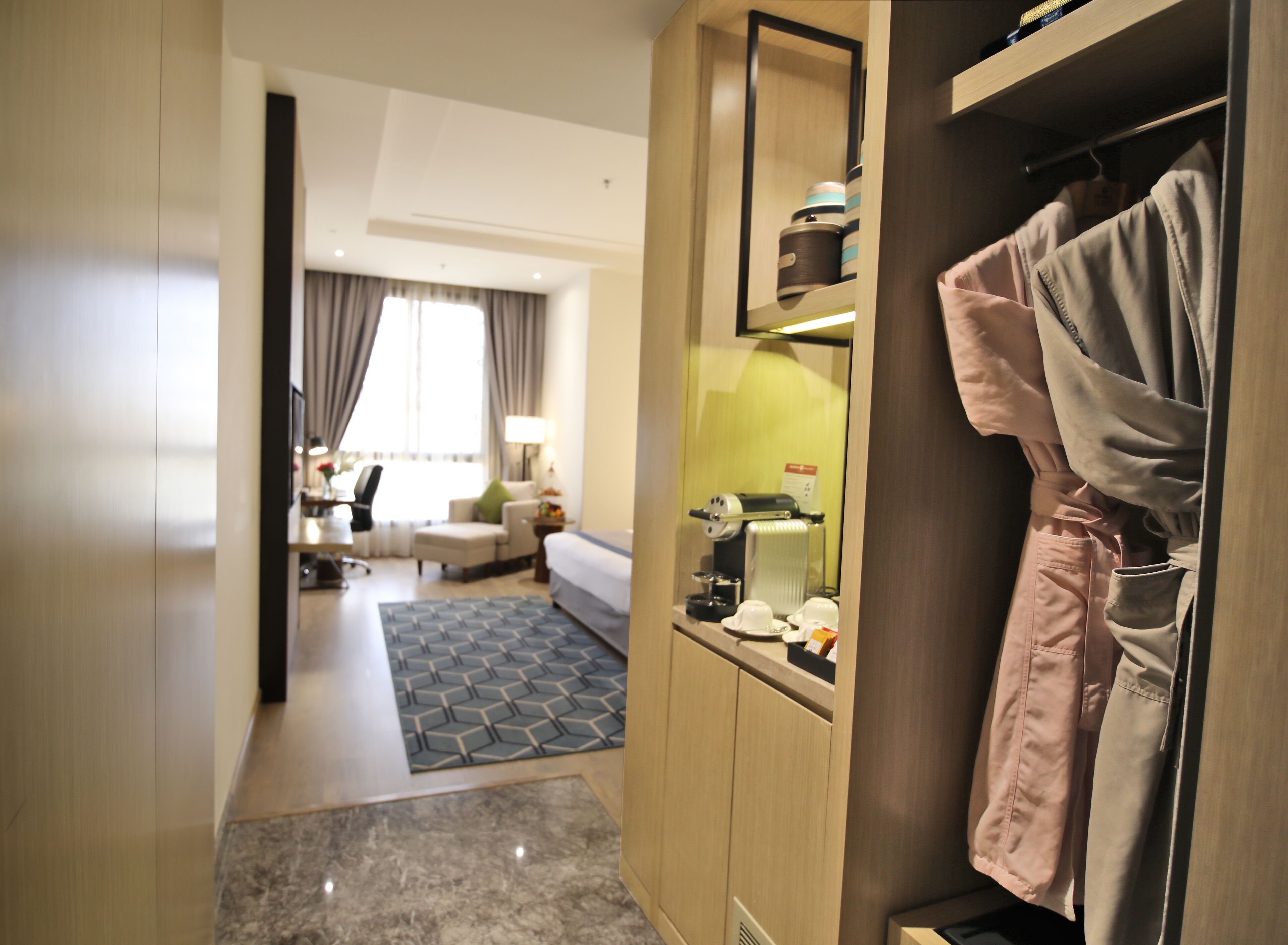 A hotel room with a bed, a seating area with a sofa and chairs, a coffee machine, and a wardrobe with clothes hanging inside.