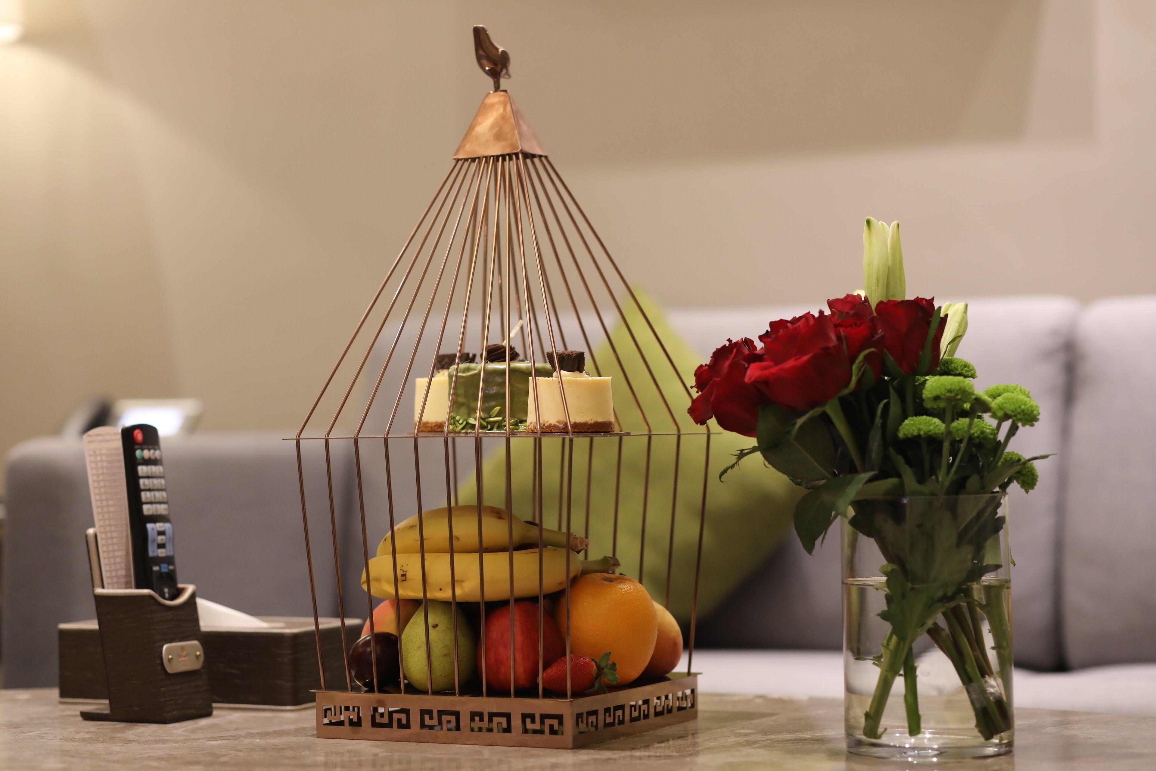A table with a fruit basket, a vase with flowers, and a remote control.