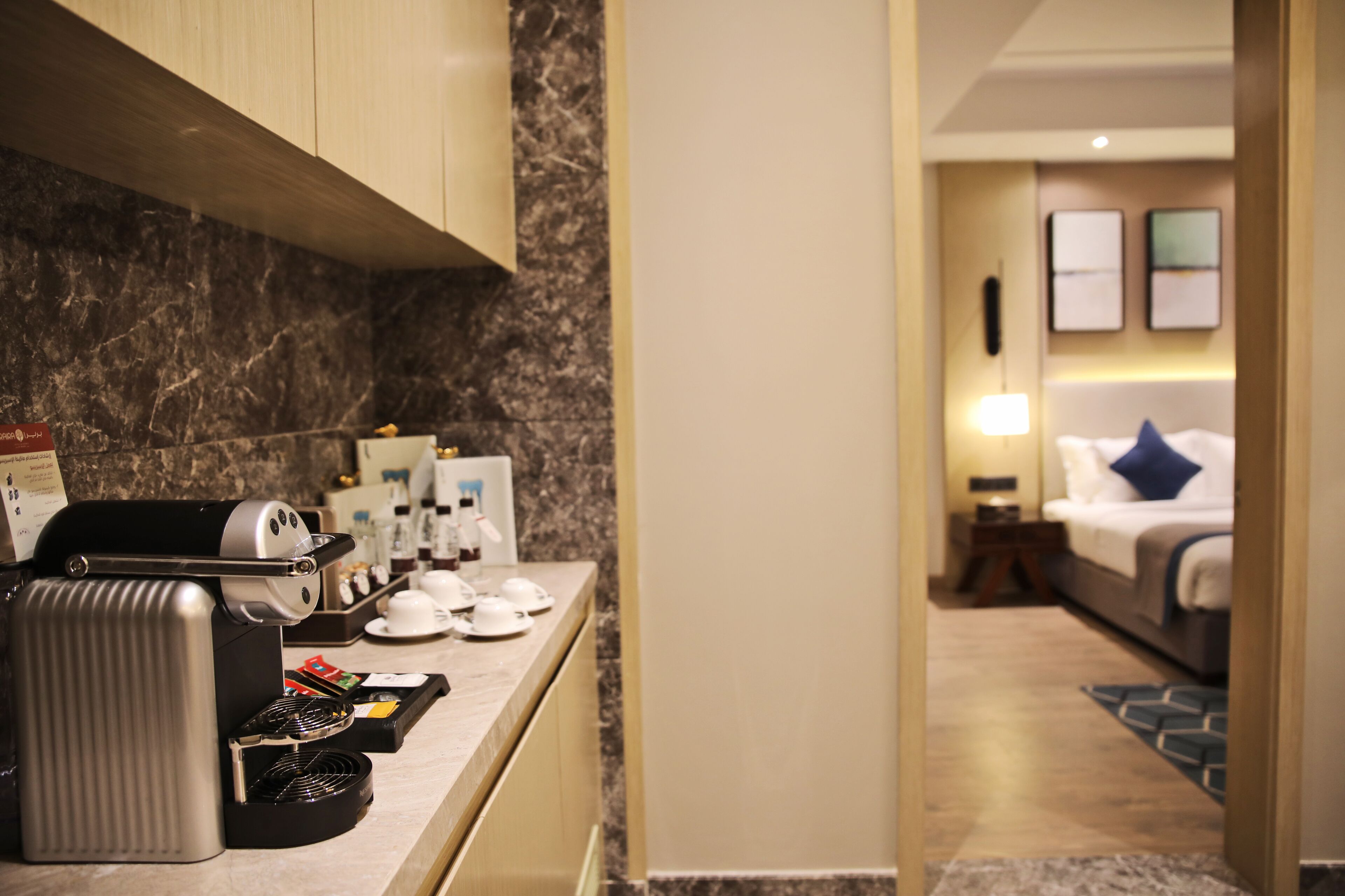 A hotel kitchen with a coffee machine, tea station, and a view into a bedroom with a bed and nightstand.