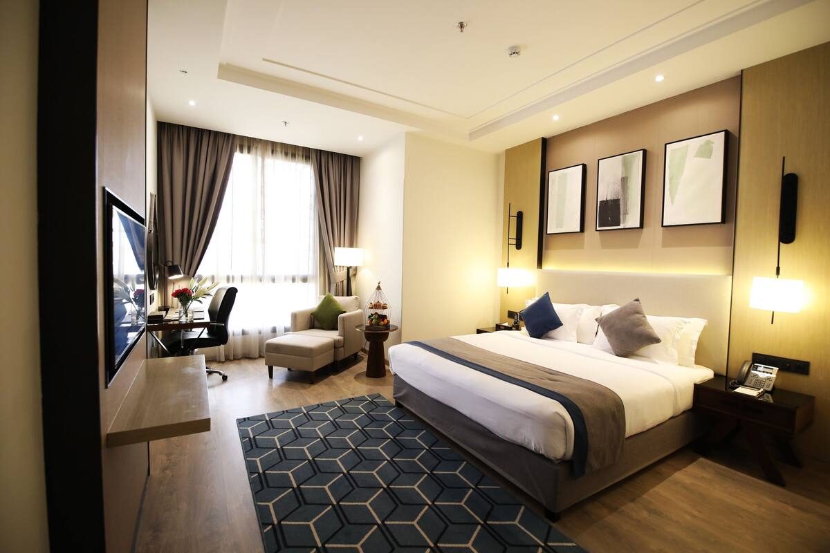 A hotel room with a large bed, a desk, a chair, a small table with a lamp, and a view of the city through the window.