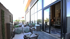 Un área de estar exterior con muebles de mimbre y cojines, una puerta de vidrio que da a un espacio interior y vistas a la calle.