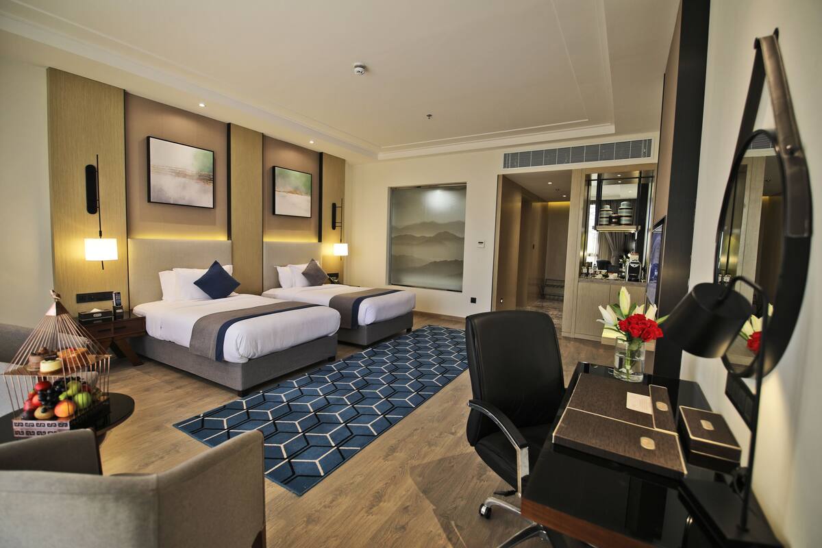 A hotel room with two beds, a desk with a mirror, a chair, a desk lamp, and a decorative vase with flowers.