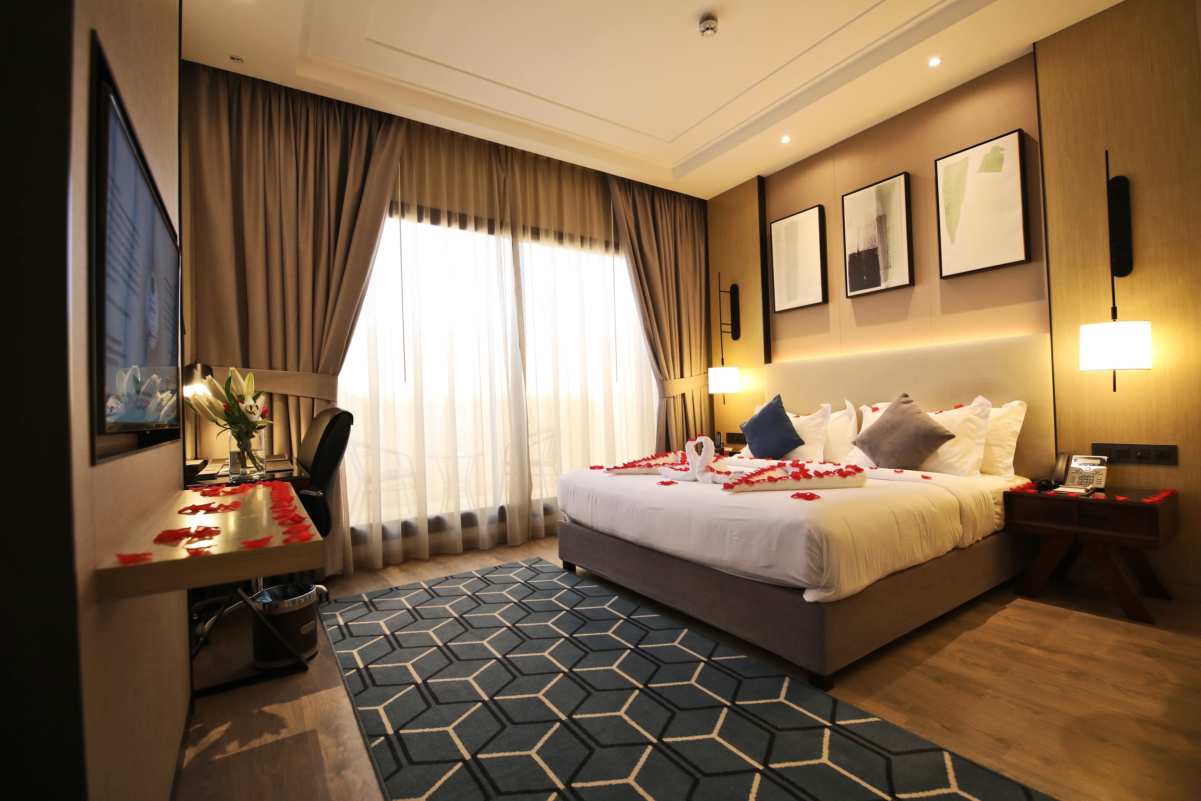A hotel room with a large bed, a desk with a computer, a chair, a desk lamp, and a bedside table with a phone.