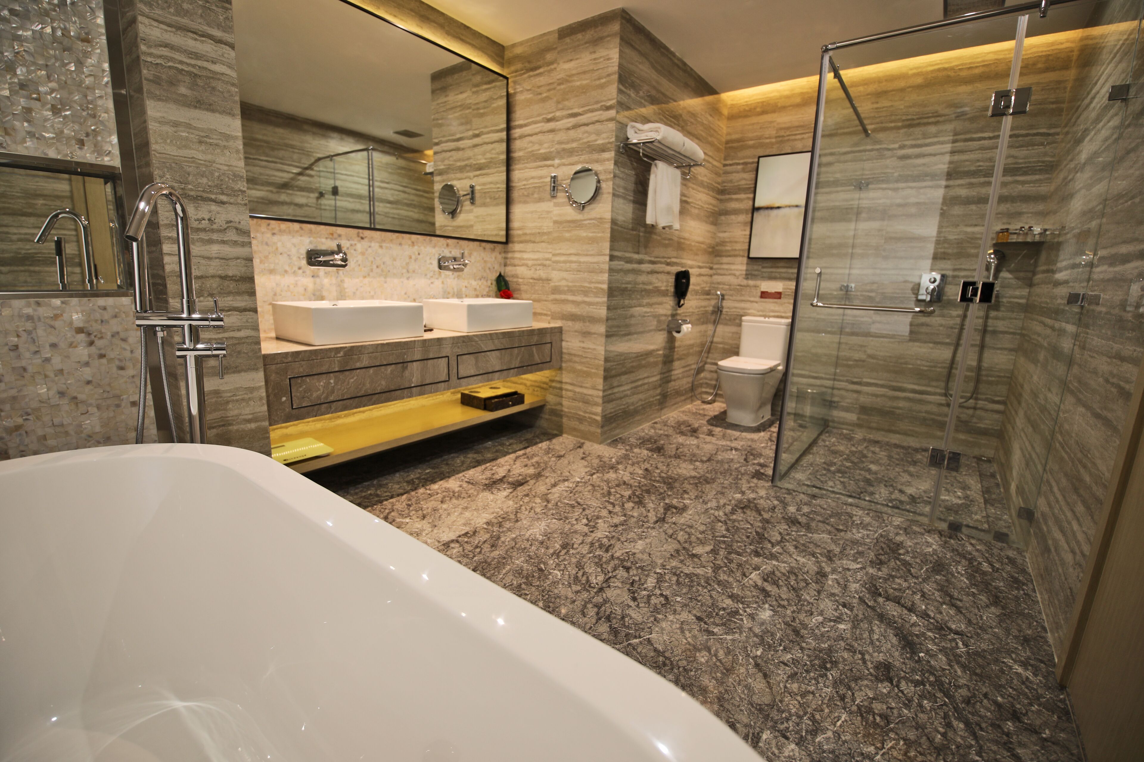 A modern bathroom with a bathtub, two sinks, a glass-enclosed shower, and marble walls.