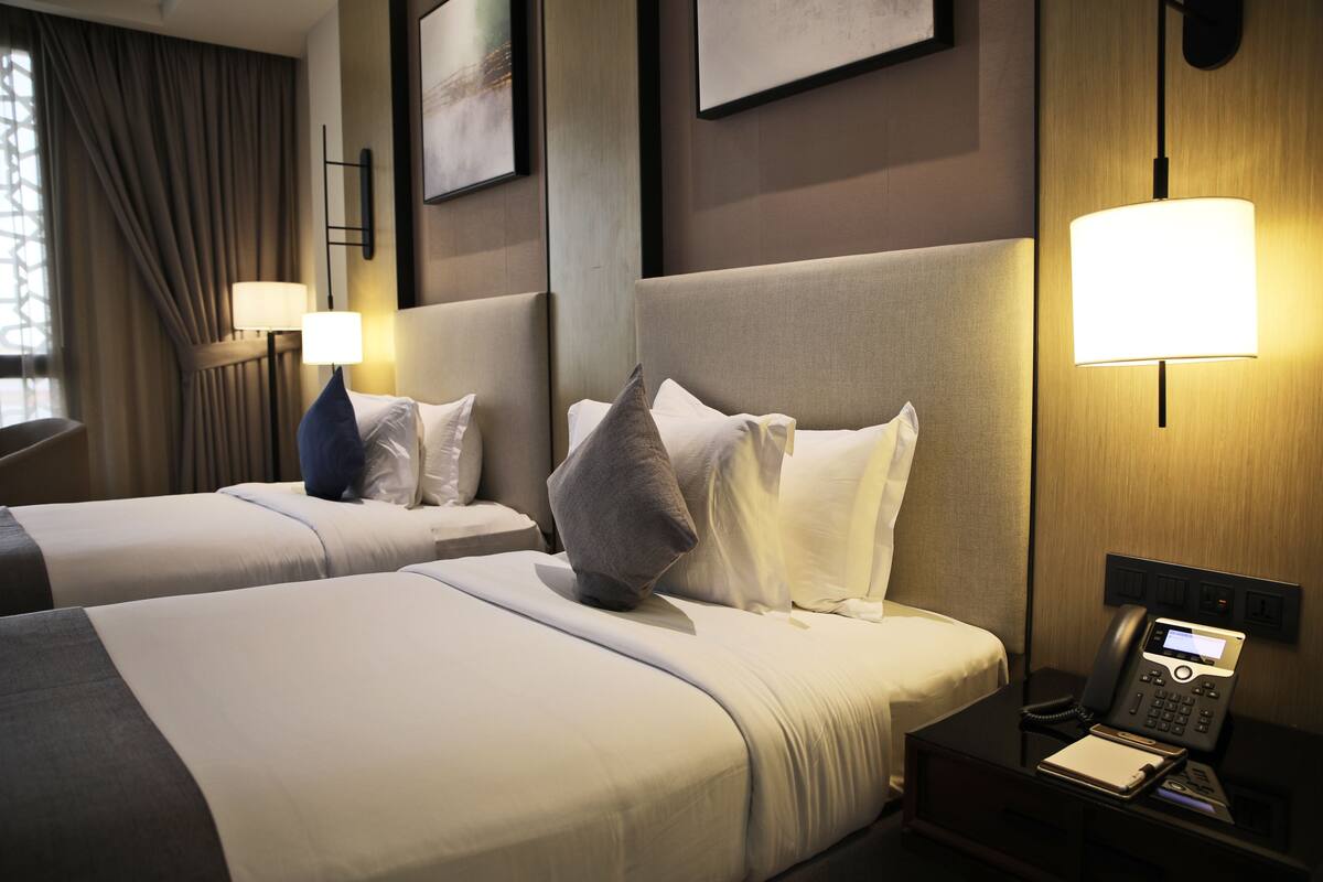 A hotel room with two beds, a bedside table with a lamp, a phone, and a window with curtains.