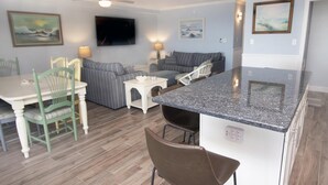 A compact living space with a dining area, a sofa, a TV mounted on the wall, and a kitchen island. - Newly renovated 2 bedroom, 2 bath condo with beautiful views of the ocean! (Ocean City)