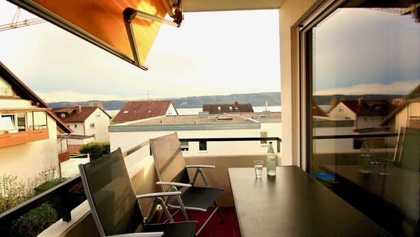 A balcony with a table and chairs, a view of houses across a body of water, and a clear sky.