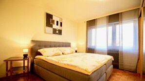 A bedroom with a bed, a bedside table, a lamp, a painting on the wall, and a window with curtains.
