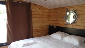 A wooden cabin room with a bed, white linens, a window with brown curtains, and a round mirror on the wall. - Toue cabanée spa (Chassenard)