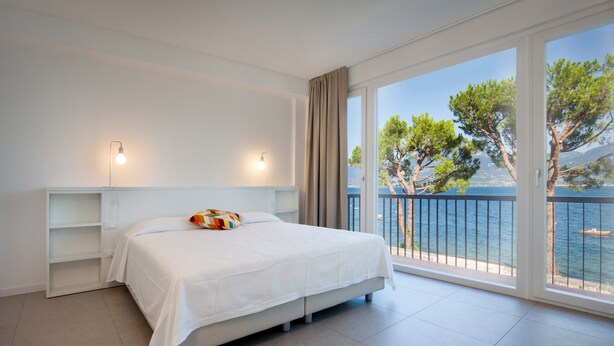 Un dormitorio con cama, dos mesitas de noche, un ventanal con vistas al mar y a árboles, y un balcón con barandilla.
