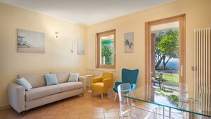 Een woonkamer met een beige bank, een gele fauteuil, een blauwe fauteuil en een glazen salontafel. Er hangen twee schilderijen aan de muur en een raam met uitzicht op groen.
