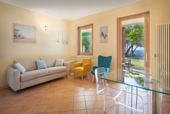 A living room with a beige sofa, a yellow armchair, a blue armchair, and a glass coffee table. There are two paintings on the walls and a window with a view of greenery. - Villetta Blue Panorama With Private Jacuzzi (Torri del Benaco)