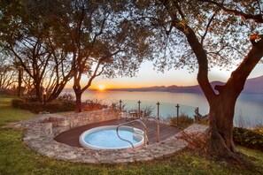 A hot tub surrounded by stone walls, with a wooden deck and metal railings, overlooking a body of water and mountains during sunset. - Villetta Blue Panorama With Private Jacuzzi (Torri del Benaco)