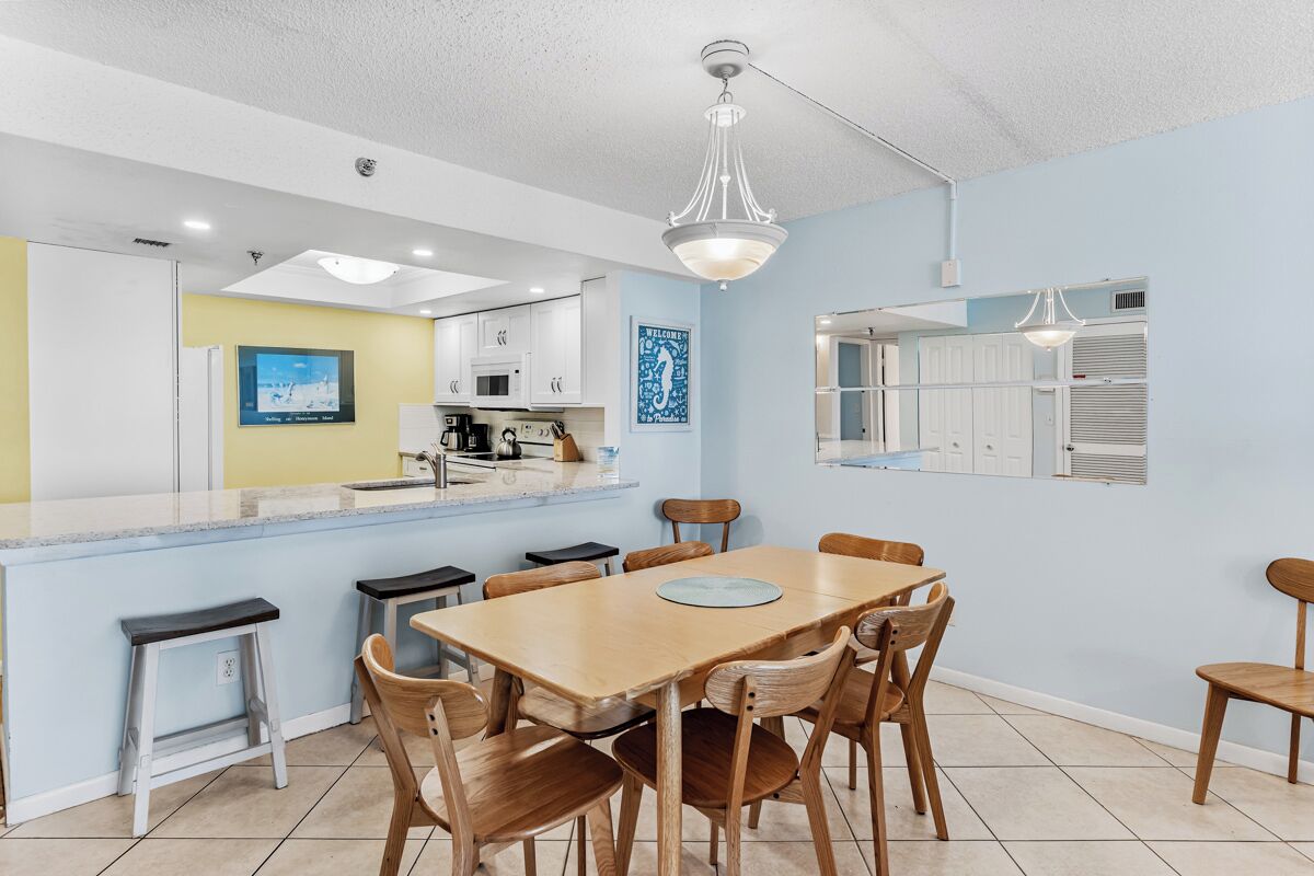 A dining area with a wooden table and chairs, a kitchen in the background, and a ceiling light fixture.