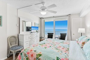 Quarto com cama, penteadeira, cadeira, escrivaninha, ventoinha no teto e varanda com vista para o mar.